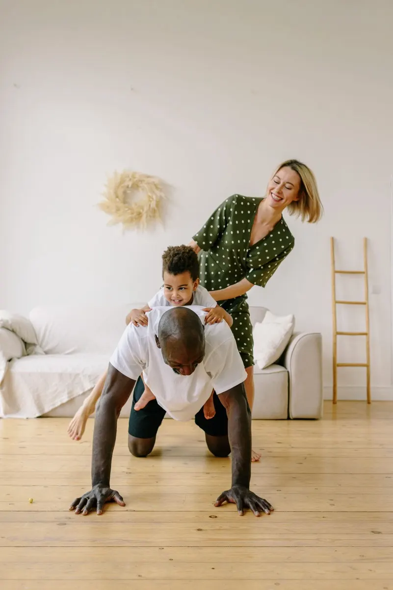 A father doing push-ups at home with his son on his back while his partner laughs